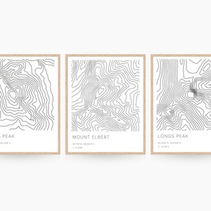 May include: Three minimalist black and white topographic maps of Pikes Peak, Mount Elbert, and Longs Peak. Each map is framed in a light brown wood frame and has the name of the mountain and its elevation and coordinates printed below the map.