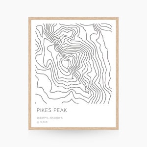 May include: A framed print featuring a black and white topographic map of Pikes Peak. The map shows contour lines and includes the mountain's name, coordinates, and elevation of 14,114 ft. The frame is made of light-colored wood.