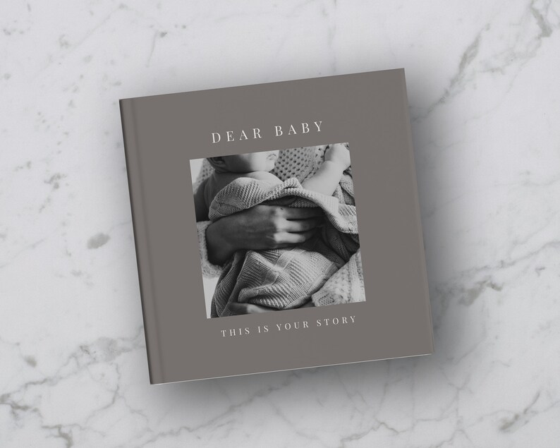 Puede incluir: Un &aacute;lbum de fotos gris con el texto "DEAR BABY THIS IS YOUR STORY" en la portada. La portada presenta una foto en blanco y negro de un beb&eacute; que est&aacute; siendo sostenido por una persona.