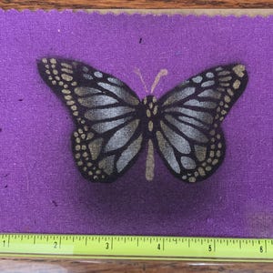 May include: A monarch butterfly design in black, silver, and gold is centered on a purple background. The butterfly is detailed with silver and gold accents. A yellow ruler is at the bottom, measuring up to 7 inches.