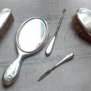 Può includere: Un set di cinque strumenti per la cura vintage argentati, tra cui uno specchio da mano, una spazzola per capelli, una lima per unghie e due strumenti per cuticole. Gli strumenti sono disposti su una superficie grigio chiaro.