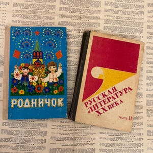 May include: Two vintage books with colorful covers rest on a background of aged newspaper pages. One book has a blue cover with a cartoon illustration and Cyrillic text. The other book has a red cover with Cyrillic text.