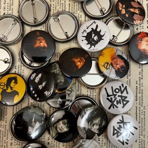 Könnte beinhalten: Eine Sammlung runder Anstecknadeln mit verschiedenen Bildern und Designs. Die Anstecknadeln sind größtenteils schwarzweiß, einige mit Farbakzenten. Einige der Bilder zeigen eine Silhouette einer Person, eine Band und ein grafisches Design.