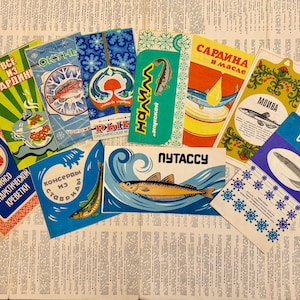Op de afbeelding: Een verzameling vintage etiketten voor sardine- en visblikken. De etiketten bevatten kleurrijke illustraties van vissen, sneeuwvlokken en andere ontwerpen. De etiketten zijn in verschillende talen en zijn gerangschikt op een achtergrond van gedrukte tekst.