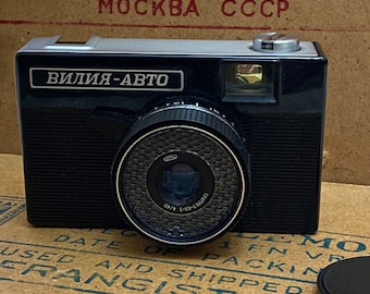 May include: A vintage black and silver "Vilia-Auto" camera with a lens and a silver top. The camera has the text "ВИЛИЯ-АВТО" on the front. The background features text in Russian and English on a cardboard surface.