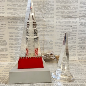 Puede incluir: Una escultura de vidrio transparente de un edificio con una base roja y una estrella roja en la parte superior. La escultura está sentada sobre una base blanca. Hay una segunda escultura de vidrio transparente más pequeña de un edificio en el fondo.