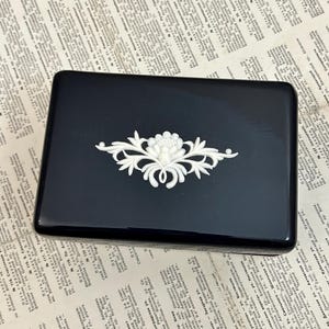 May include: A black rectangular box with a white floral design on the lid. The design features a stylized flower with leaves and a pearl-like center.