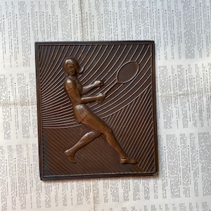 May include: A bronze wall plaque depicting a woman playing tennis. The plaque has a textured surface and a raised design of the woman holding a tennis racket.