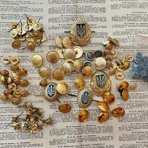May include: A collection of gold and blue buttons with a trident symbol, a Ukrainian national symbol. The buttons are arranged on a white surface with text in a foreign language. There are also some small gold stars and a bag of blue buttons.