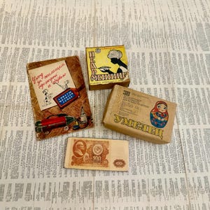 May include: A collection of vintage items, including a small box with a woman's silhouette, a wooden box with a Matryoshka doll illustration, a postcard with text, and a 100-ruble bill, all arranged on a newspaper background.