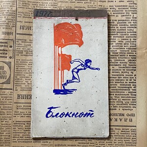 May include: A vintage, red, white, and blue notebook with a drawing of a woman jumping over a red flag. The notebook has the word "Блокнот" written in blue ink.