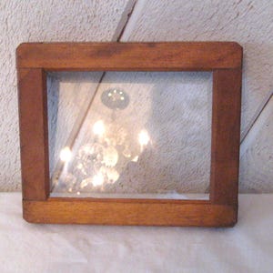 May include: A rectangular wooden frame with a clear glass insert. The frame is made of a medium-toned wood and has a simple, classic design. The glass reflects light, creating a bright and airy feel. The frame is likely for a picture or artwork.