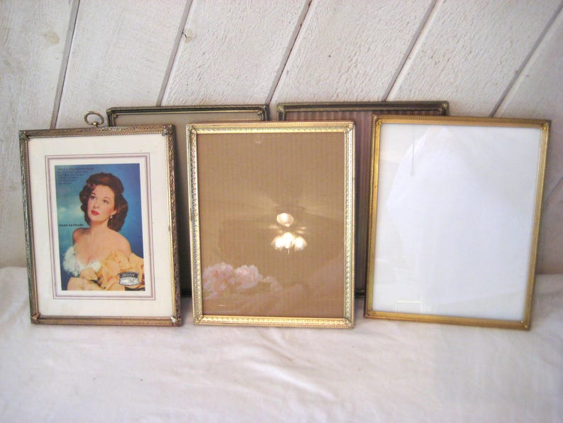 Puede incluir: Una colecci&oacute;n de marcos de fotos antiguos en varios tama&ntilde;os y estilos. Los marcos tienen acabados dorados y plateados, algunos con detalles ornamentados. Un marco contiene un anuncio vintage con una mujer. Los marcos est&aacute;n dispuestos sobre una superficie blanca.
