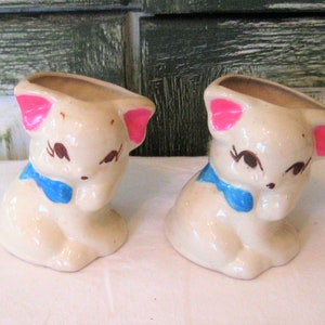 May include: Two ceramic kitten planter pots with blue and pink painted details. The kittens are sitting with their paws tucked in and have closed eyes.