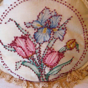 Könnte beinhalten: Ein weißes Stoffkissen mit einer Spitzenborte zeigt ein gesticktes Design aus rosa Tulpen und einer blauen Iris. Die Stickerei ist mit kleinen roten Perlen umrandet.