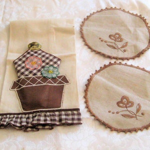 May include: A set of three vintage kitchen linens featuring a brown embroidered flower pot with flowers and a brown gingham border. The set includes two round doilies with embroidered flowers.