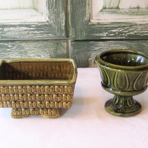 May include: Two green ceramic planters. One is a rectangular planter with a textured surface and four legs. The other is a round planter with a pedestal base and a textured surface.