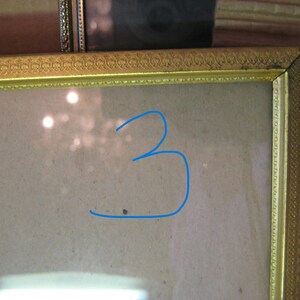 Puede incluir: Un marco de fotos dorado con detalles ornamentados. El marco rodea una superficie de color claro, con el n&uacute;mero "3" dibujado en azul en la superficie. El marco es rectangular y parece ser vintage.