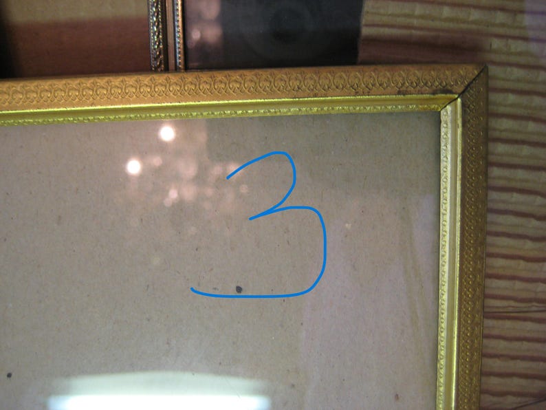 Puede incluir: Un marco de fotos dorado con detalles ornamentados. El marco rodea una superficie de color claro, con el n&uacute;mero "3" dibujado en azul en la superficie. El marco es rectangular y parece ser vintage.