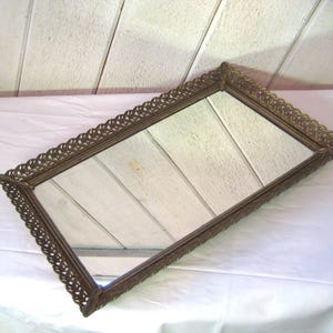 May include: An antique rectangular vanity tray with a mirrored surface and a decorative, ornate bronze-colored frame. The frame features a detailed, repeating pattern around the edges. The tray is resting on a white surface.