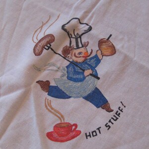 May include: A white tea towel with a hand-painted design of a chef carrying a sausage, a donut, and a cup of coffee. The chef is wearing a white chef's hat and a blue and white uniform. The text "HOT STUFF!" is written in black below the chef.