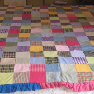 May include: A patchwork quilt with a variety of colors and patterns, including plaid, stripes, and solids. The quilt has a blue ruffle around the edge and a pink ruffle around the bottom.