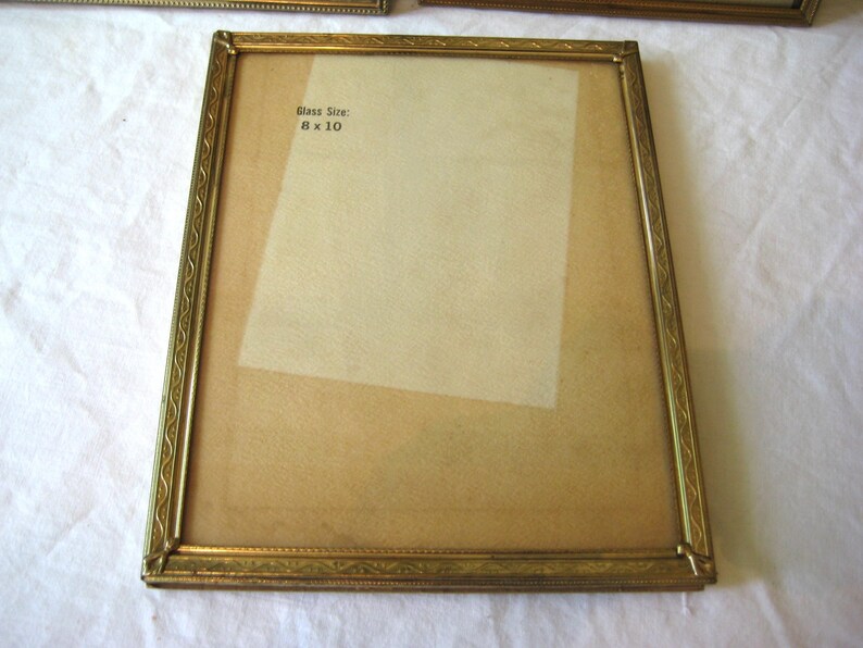 Puede incluir: Un marco de fotos rectangular dorado con un borde ornamentado. El marco tiene un tama&ntilde;o de vidrio de 8 x 10 pulgadas (20 x 25 cm). El marco est&aacute; vac&iacute;o, con un respaldo de color claro visible a trav&eacute;s del vidrio.