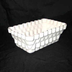 May include: A white rectangular ceramic planter with a textured, woven-like pattern. The planter has a scalloped rim and is set against a dark background. This decorative piece is suitable for indoor or outdoor use.