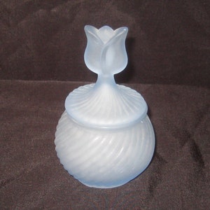 Könnte beinhalten: Eine gefrostete Glasdose mit einem wirbelnden Design und einem bläulich getönten Glasdeckel in Form einer Blume.