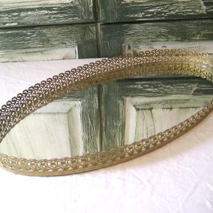 May include: Oval vintage vanity tray with a mirrored surface and a decorative filigree metal edge. The gold-toned tray sits on a white surface. The tray measures approximately 46 cm long and 25 cm wide.