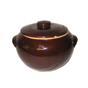 Può includere: Una pentola in ceramica marrone scuro con coperchio abbinato. La pentola ha due piccole maniglie su entrambi i lati e un piccolo pomello sul coperchio. La pentola è rotonda e ha una finitura lucida. Probabilmente viene utilizzata per cucinare o conservare.