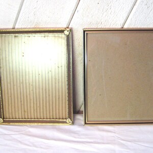 May include: Two rectangular picture frames with gold-colored metal frames. One frame has a textured, ribbed backing, while the other has a plain, light brown backing. The frames are displayed on a white surface with a light-colored wooden background.