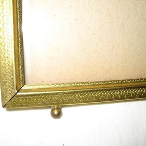 Puede incluir: Primer plano de un marco de fotos dorado con detalles ornamentados. El marco tiene un patr&oacute;n decorativo a lo largo de los bordes y un peque&ntilde;o pie redondo. El marco est&aacute; dise&ntilde;ado para contener una fotograf&iacute;a o una obra de arte.