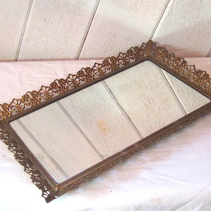 May include: A gold-colored metal tray with a mirrored surface and a decorative, ornate border. The tray is rectangular and has a delicate, floral design.
