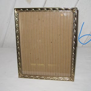 Puede incluir: Un marco de fotos rectangular dorado con un borde decorativo. El marco tiene una parte delantera de vidrio y una parte trasera de cart&oacute;n. El marco est&aacute; vac&iacute;o y listo para exhibir una fotograf&iacute;a o una obra de arte.