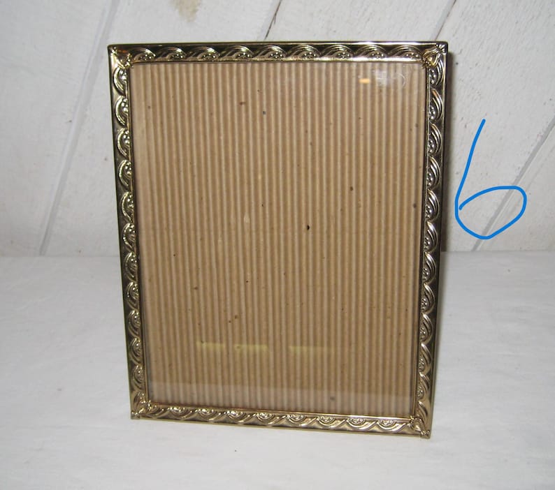 Puede incluir: Un marco de fotos rectangular dorado con un borde decorativo. El marco tiene una parte delantera de vidrio y una parte trasera de cart&oacute;n. El marco est&aacute; vac&iacute;o y listo para exhibir una fotograf&iacute;a o una obra de arte.