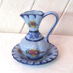 May include: A light blue ceramic pitcher and matching plate set. The pitcher has a flared top and a curved handle. Both pieces are decorated with floral designs in red, yellow, and green, outlined in dark blue.