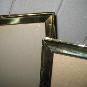 Puede incluir: Dos marcos dorados con forma rectangular. Los marcos tienen un acabado met&aacute;lico y est&aacute;n colocados en &aacute;ngulo, mostrando sus esquinas y bordes. El fondo es una superficie de madera de color claro.