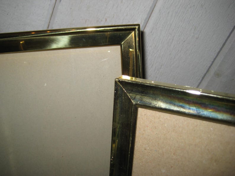 Puede incluir: Dos marcos dorados con forma rectangular. Los marcos tienen un acabado met&aacute;lico y est&aacute;n colocados en &aacute;ngulo, mostrando sus esquinas y bordes. El fondo es una superficie de madera de color claro.