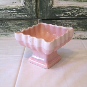 Könnte beinhalten: Ein quadratischer, rosa-weißer Keramik-Blumentopf im Vintage-Stil mit gewelltem Rand. Der Topf hat einen Sockel und eine glänzende Oberfläche. Das Farbschema ist ein zartes Rosa mit weißen Akzenten.