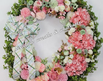 May include: A pink and green floral wreath with a plaid bow. The wreath is made of artificial flowers and greenery. The text "dixieland wreaths & whatknots" is visible on the wreath.