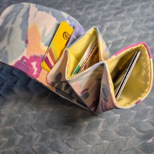 May include: A fabric accordion-style wallet featuring a pastel floral pattern in shades of pink, blue, and yellow. The interior is lined with bright yellow fabric and contains multiple slots holding several credit cards and identification cards.
