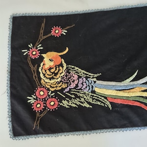 May include: A black rectangular fabric runner featuring an embroidered bird with a yellow, orange, and purple body and long, multi-colored tail feathers. The bird is perched on a brown branch with pink flowers. The edges have a light blue trim.