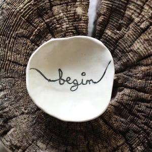 May include: A small, white ceramic dish with the word "begin" written in black script, resting on a weathered, dark brown wooden slice. The wood has visible growth rings and a rough texture. The dish is centered on the wood.