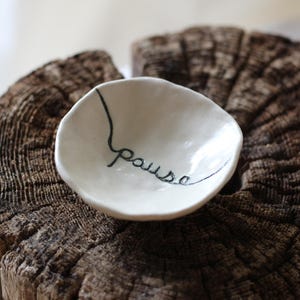 May include: A small, off-white ceramic dish with the word "pause" written in black cursive script. The dish has an organic shape and sits on a weathered, dark brown wooden surface. The overall aesthetic is rustic and minimalist.