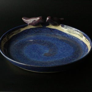 May include: A handmade ceramic bowl with a deep blue interior and a cream-colored rim. Two small, dark purple bird figures are perched on the rim. The bowl has a spiral design in the center and feather details around the edge.