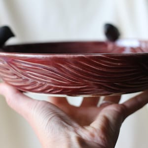 Puede incluir: Un cuenco de cerámica burdeos con un patrón texturizado en forma de olas. Dos pequeñas figuras de pájaros negros están posadas en el borde. El cuenco se sostiene en una mano, mostrando su diseño y artesanía. Una pieza decorativa.