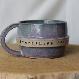 May include: A handmade ceramic mug with a purple and green glaze. The mug features a band with the words "Everything you" stamped on it. The mug is sitting on a wooden surface.