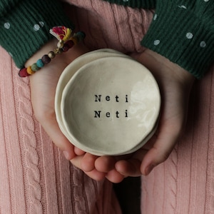 May include: A set of two white ceramic ring dishes with the words "Neti Neti" written in black. The dishes are stacked on top of each other and are being held by a person's hands.