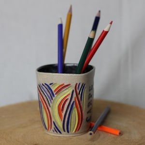 Puede incluir: Un portalápices de cerámica con base beige e interior negro. El soporte está decorado con líneas curvas y coloridas en rojo, amarillo, azul y naranja. Varios lápices de colores están dentro, con algunos más sobre la superficie de madera.